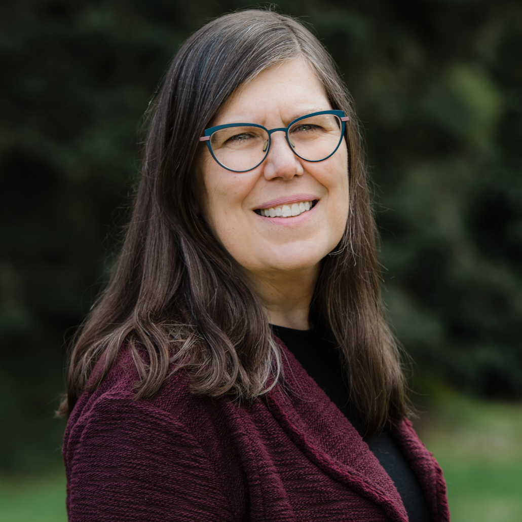 A woman with shoulder-length straight brown hair, glasses, and a burgundy sweater, smiling in an outdoor setting with greenery.