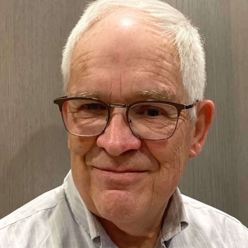 An older man with short white hair and glasses, smiling and wearing a light gray shirt.