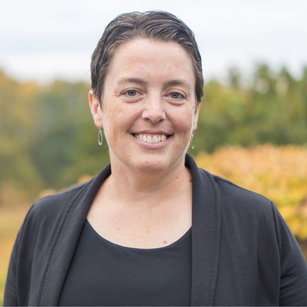 A woman with short dark hair, smiling, wearing a black top and standing in an outdoor setting with autumn foliage.