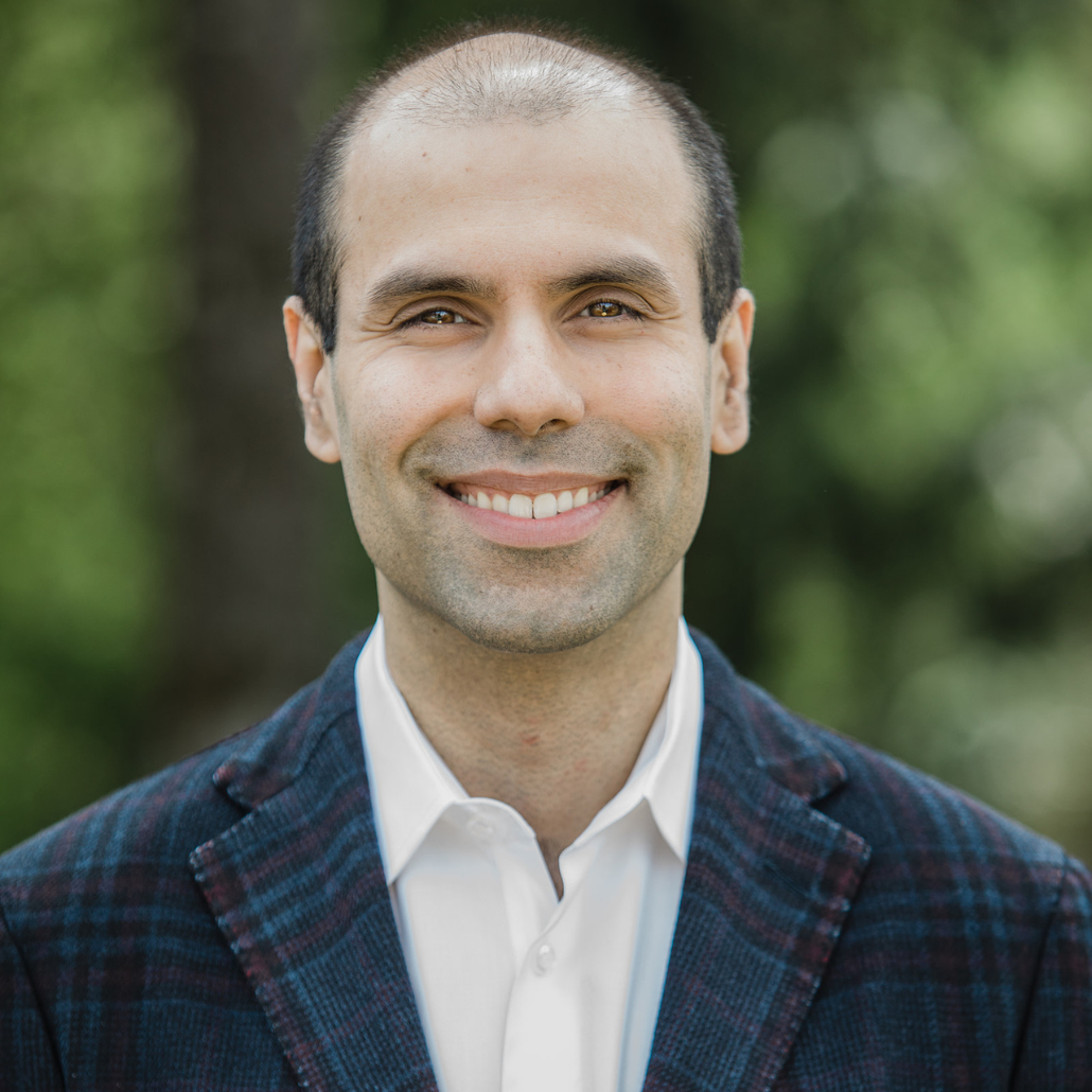 A man with short black hair, smiling, wearing a checkered suit jacket and a white shirt, standing in front of a blurred green background.