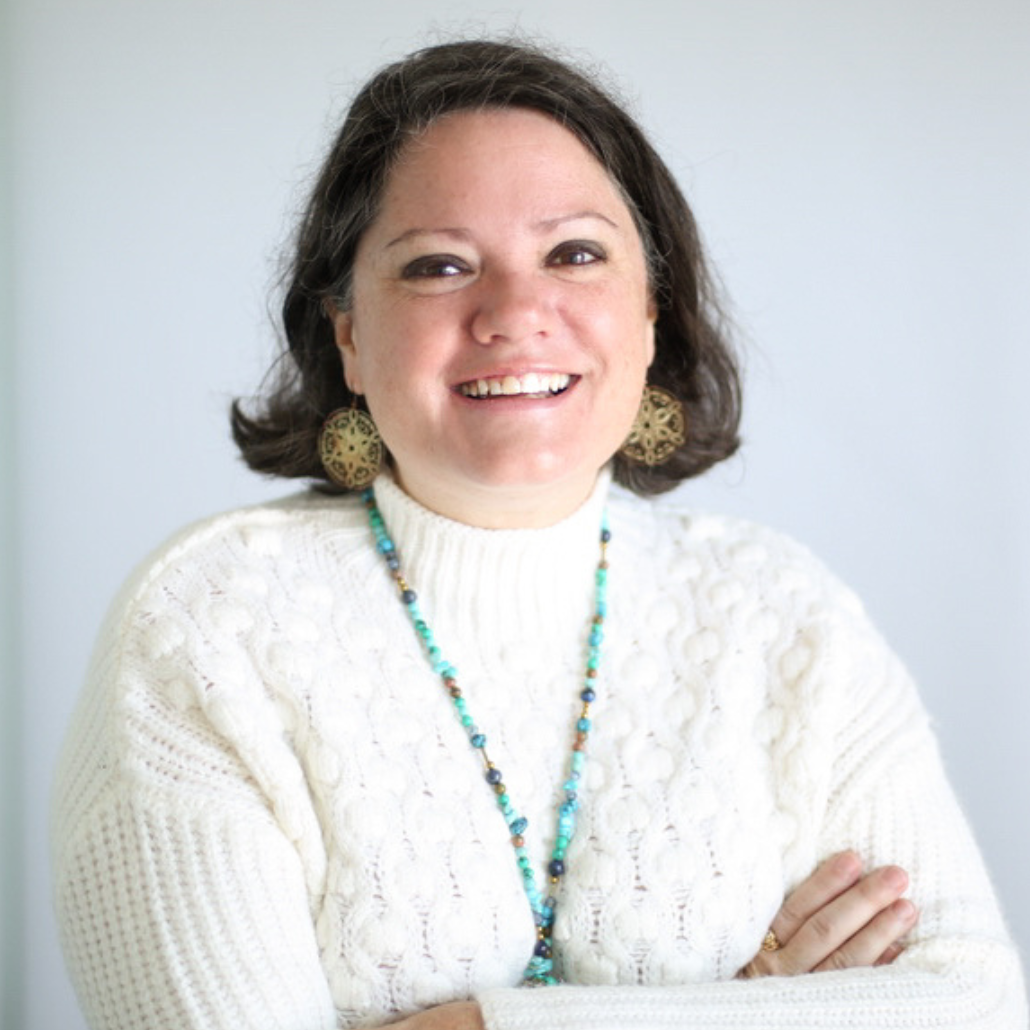 A woman with short dark hair, smiling, wearing a white sweater and turquoise necklace, standing in front of a white background.