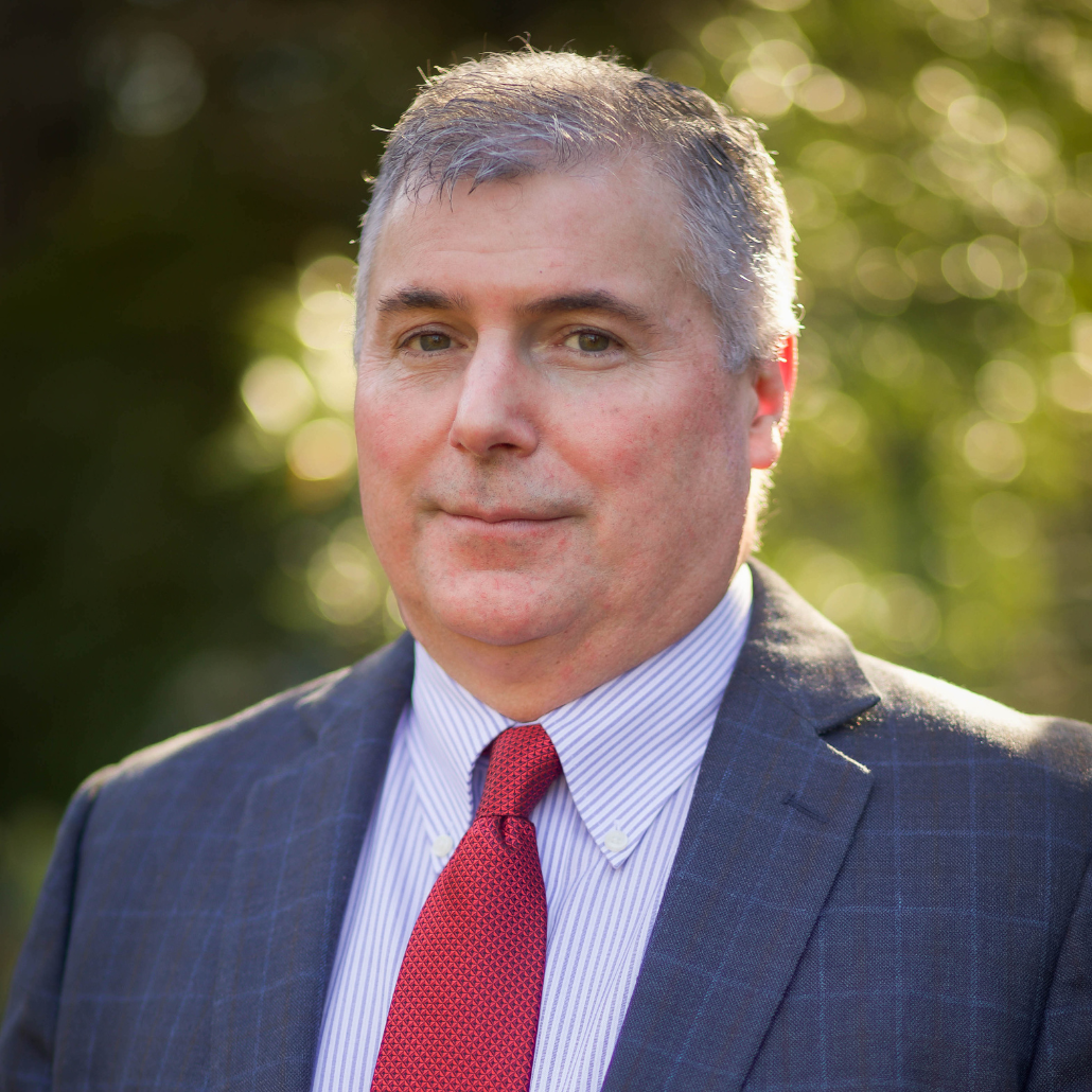 A man with short gray hair, wearing a suit, white shirt, and red tie, standing in front of a blurred natural background with bokeh effect.