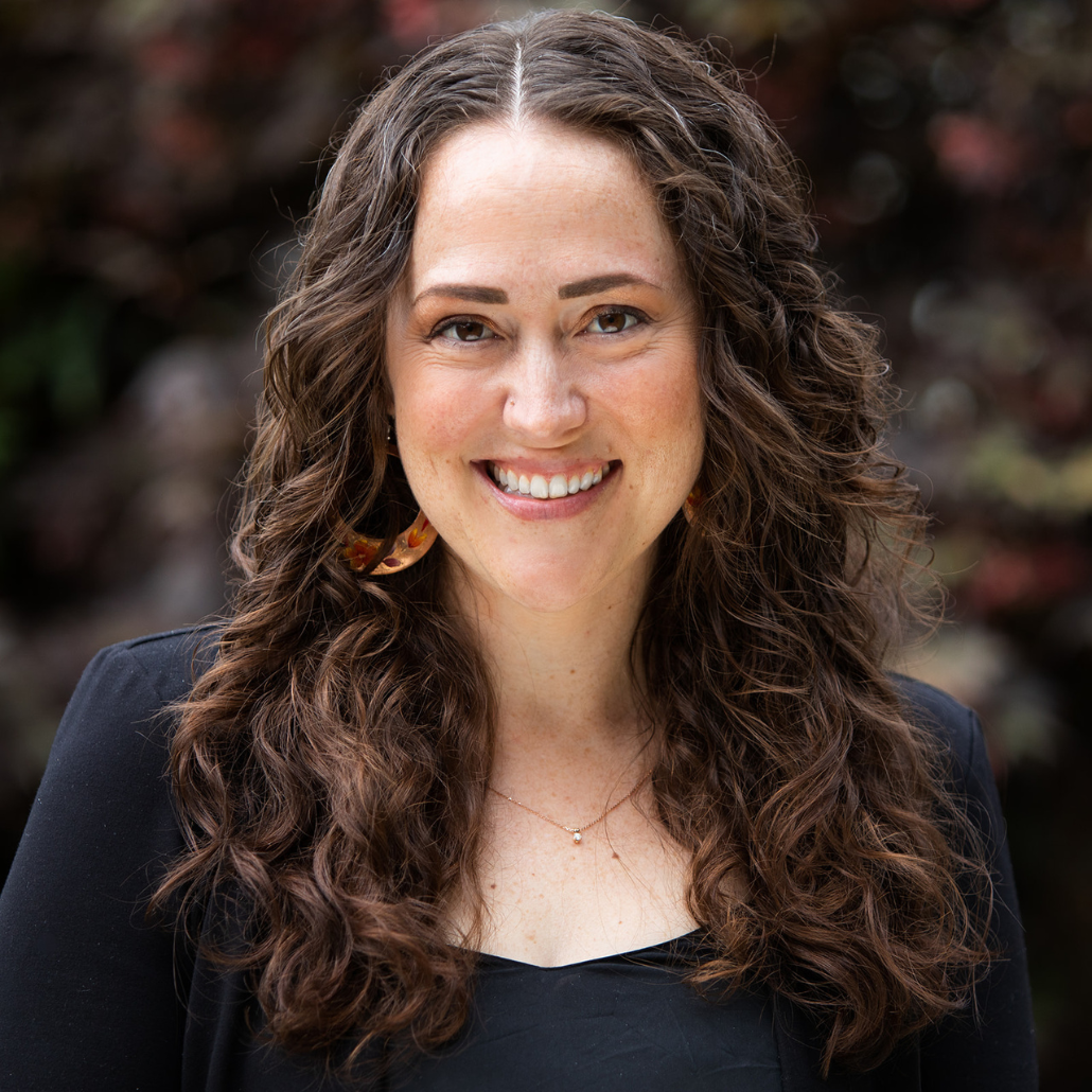 A woman with long curly brown hair, smiling, wearing a black top, standing in front of a background with colorful foliage.