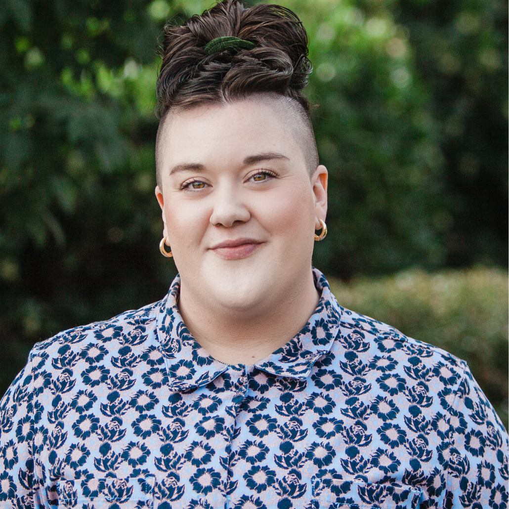 A person with short dark hair styled in a top knot, wearing gold hoop earrings and a floral patterned shirt, smiling in front of a green leafy background.