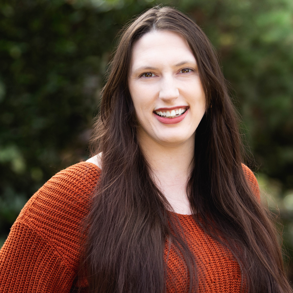 A woman with long dark brown hair, smiling, wearing a rust-colored sweater, standing in front of a green leafy background.