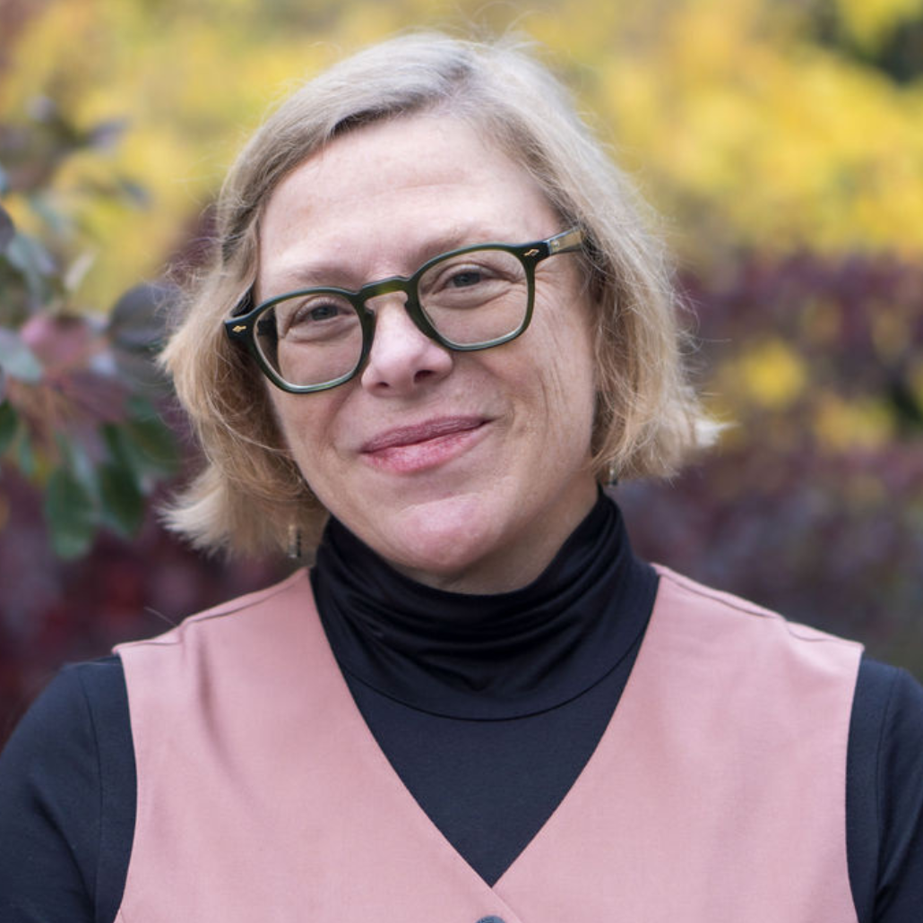 A woman with short blonde hair, wearing green-framed glasses, a black turtleneck, and a pink vest, smiling in front of a blurred background with autumn foliage.