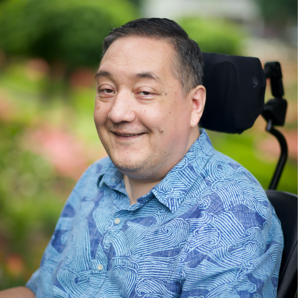  A man with short dark hair, smiling, wearing a blue patterned shirt, seated in a wheelchair in an outdoor setting with greenery and flowers in the background.