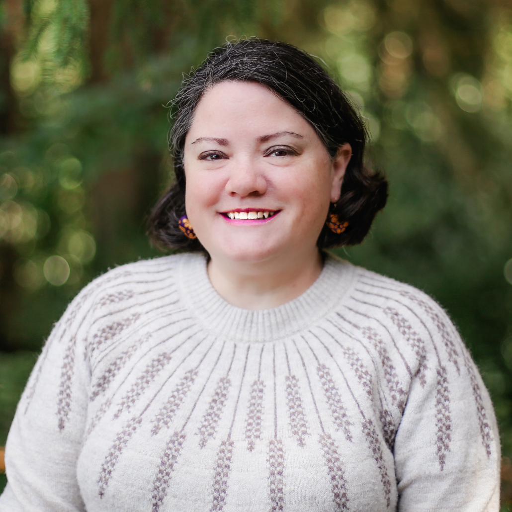 A woman with short dark hair, smiling, wearing a cream-colored sweater, and standing in front of a green natural background.