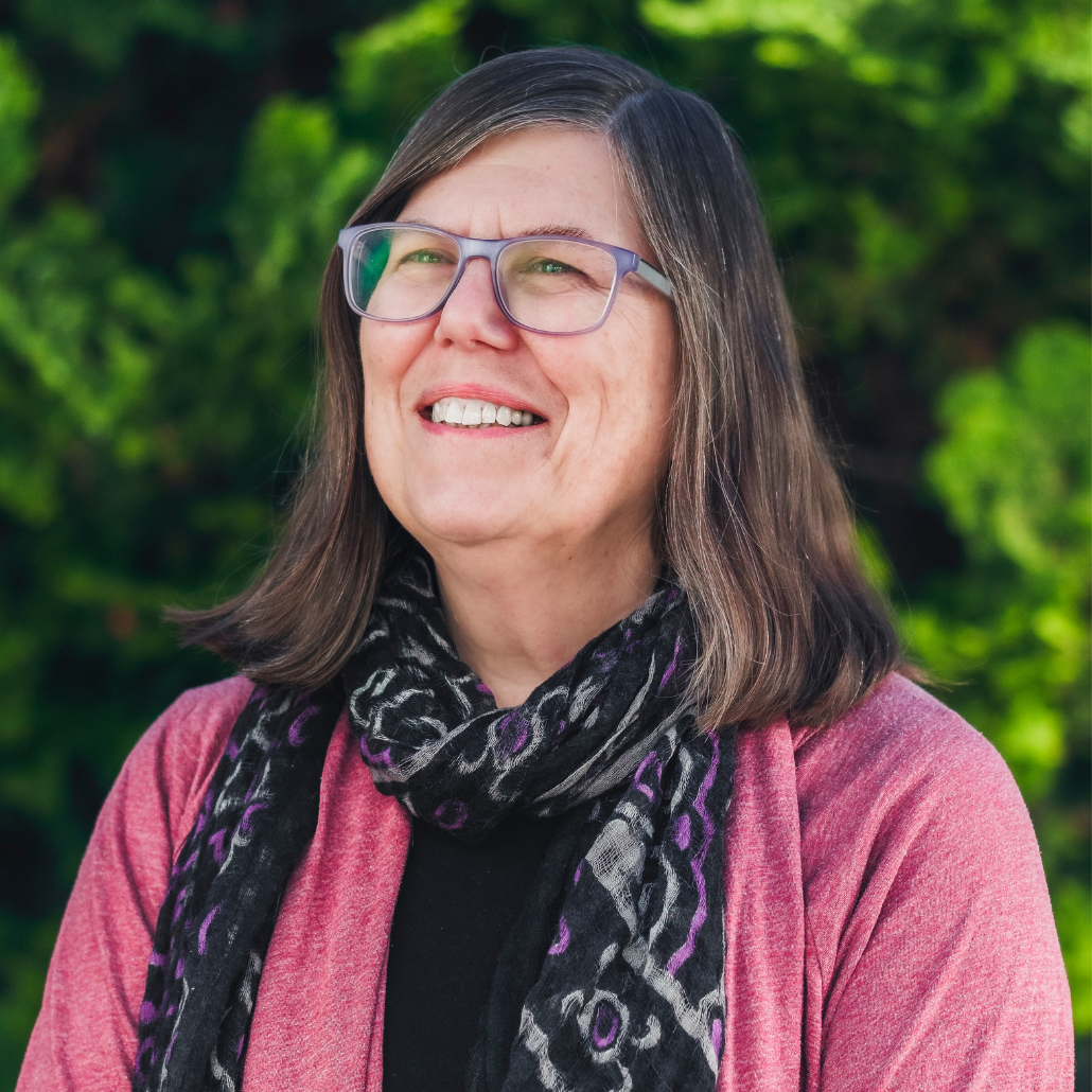 A woman with shoulder-length straight brown hair, glasses, and a pink sweater with a black and purple scarf, smiling in an outdoor setting with greenery.