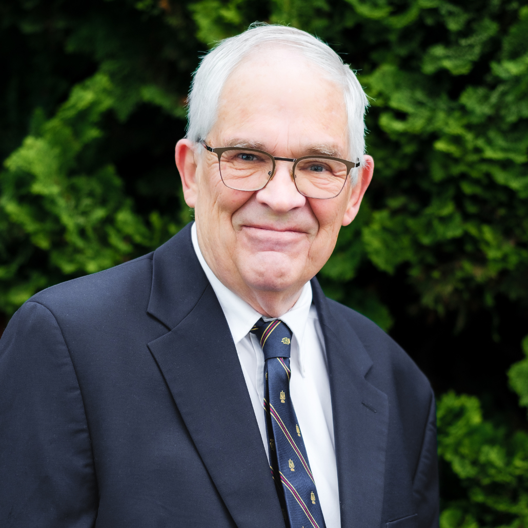 An older man with short white hair and glasses, smiling and wearing a dark navy suit, standing outdoors with trees in the background.