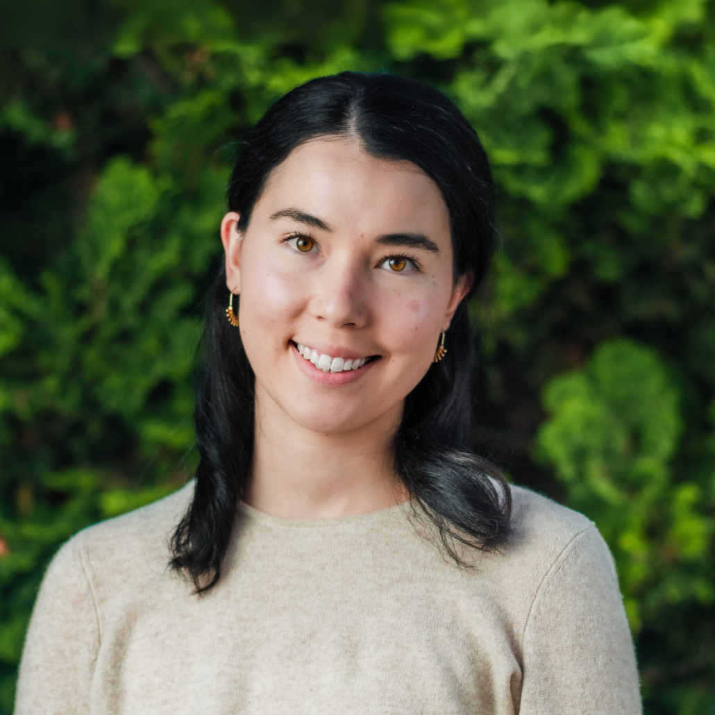 A woman with black hair, wearing a cream-colored sweater and gold earrings, smailing in an outdoor setting with a blurred background of trees and bushes. 