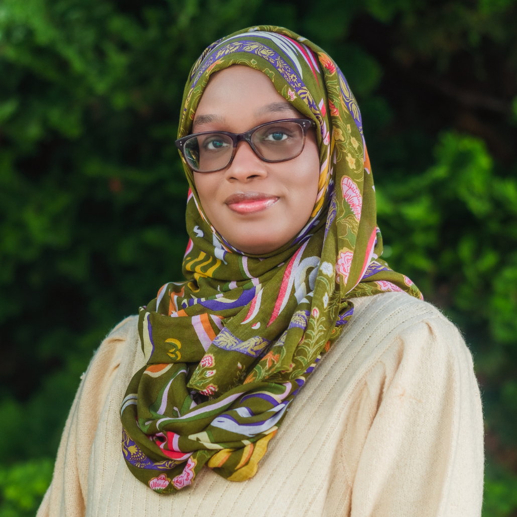 A woman with a yellow sweater and a green, purple, and pink scarf, wearing glasses, smiling in an outdoor setting with a blurred background of trees and bushes. 