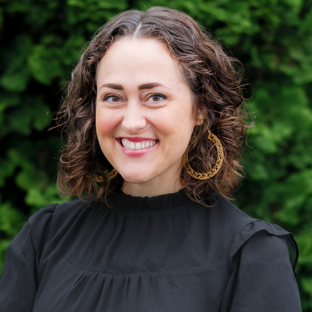 A woman with shoulder-length curly brown hair, smiling, wearing a black top and ghold earrings, standing in front of a background with dark green foliage.