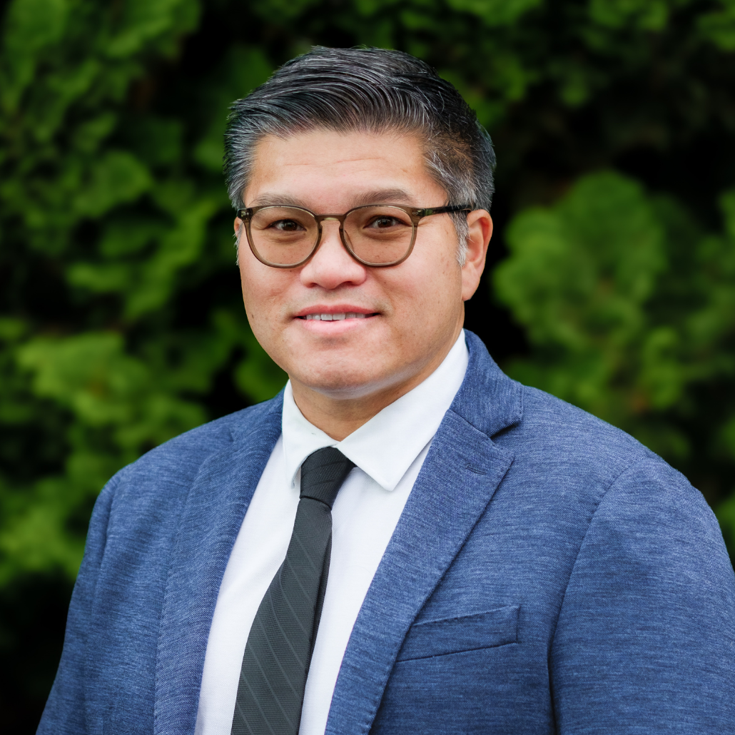 A man with short dark hair, glasses, and a blue blazer, smiling and wearing a white shirt, standing in an outdoor setting with a blurred background of trees and bushes.