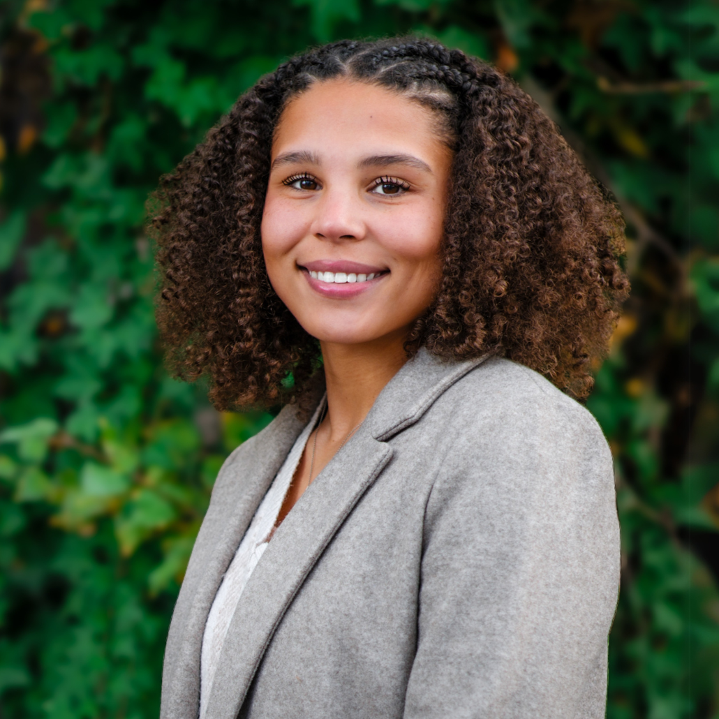 A woman smiling with curly brown hair wearing a light gray blazer and a white shirt standing in front of dark green foliage.