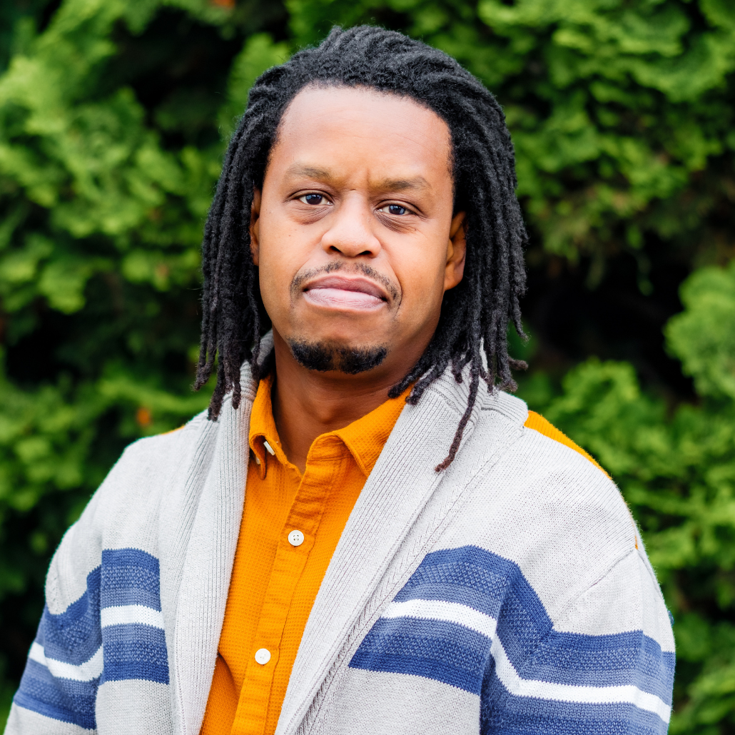 A man with dark brown dreadlocks, wearing a blue and white cardigan over an orange shirt, smiling, in an outdoor setting with a blurred background of trees and bushes. 