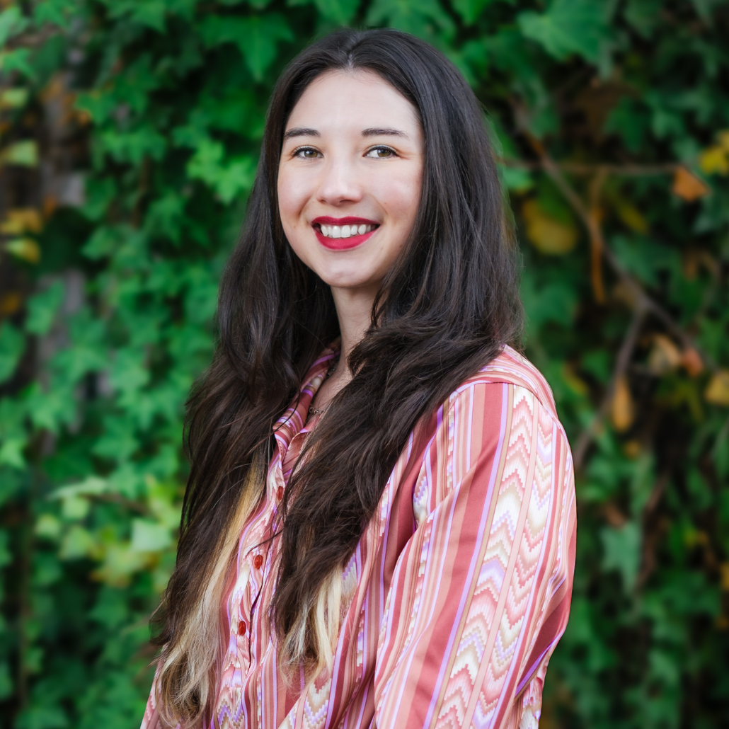 A woman smiling with long dark brown hair and a pink blouse standing in front of dark green foliage.