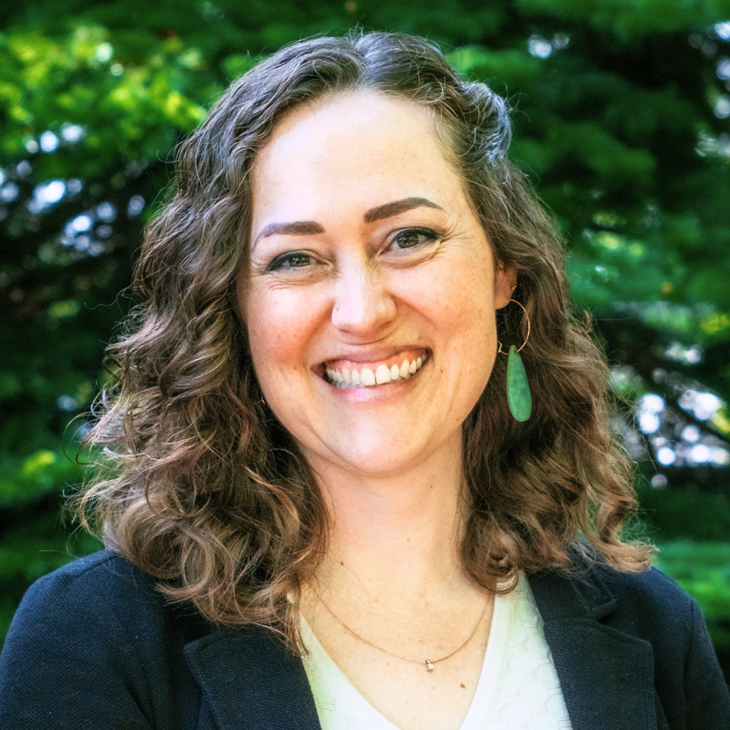 A woman with shoulder-length curly brown hair, smiling, wearing a black jacket and earrings, standing in front of a background with dark green foliage.