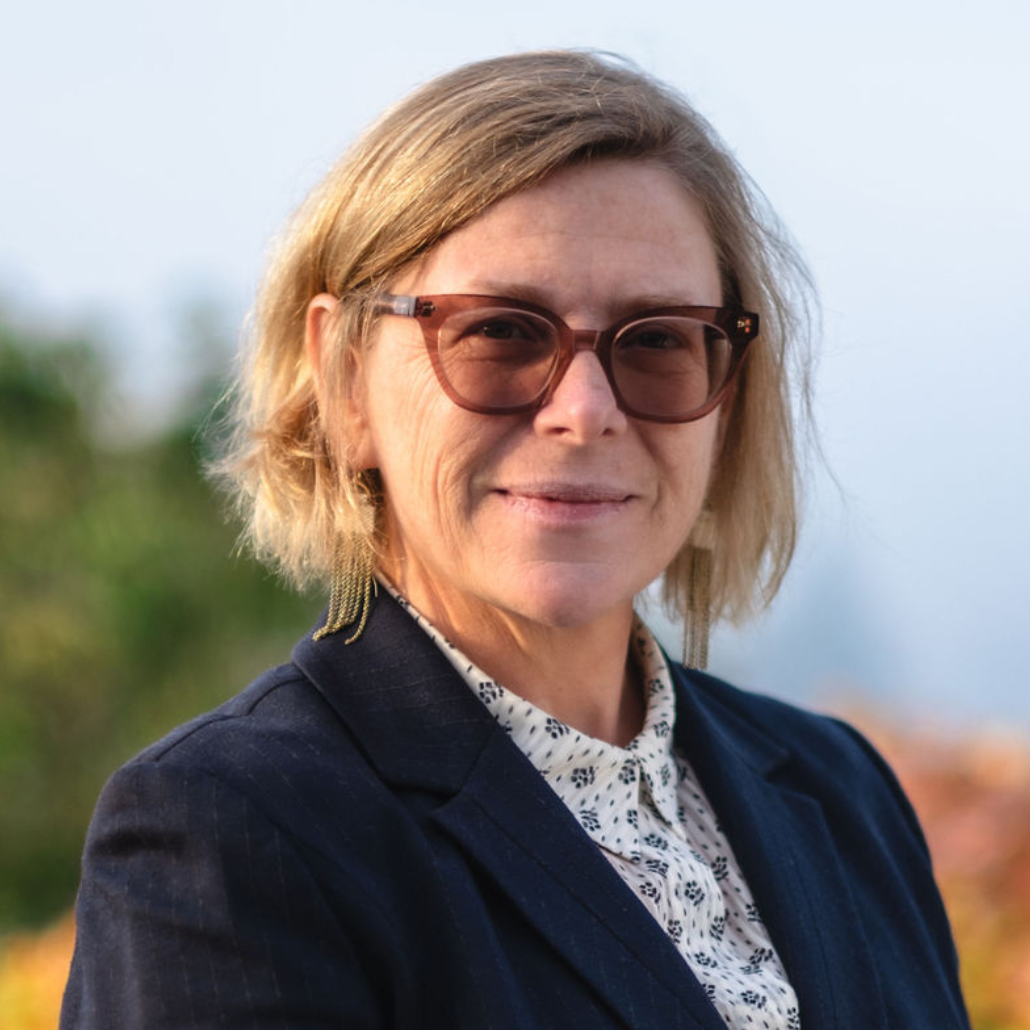 A woman with short blonde hair and wearing glasses in front of a blurred background with autumn foliage.