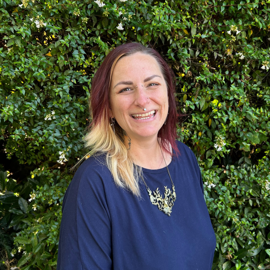 A person with red and blonde hair wearing a blue shirt and a gold necklace smiling in front of a greed hedge