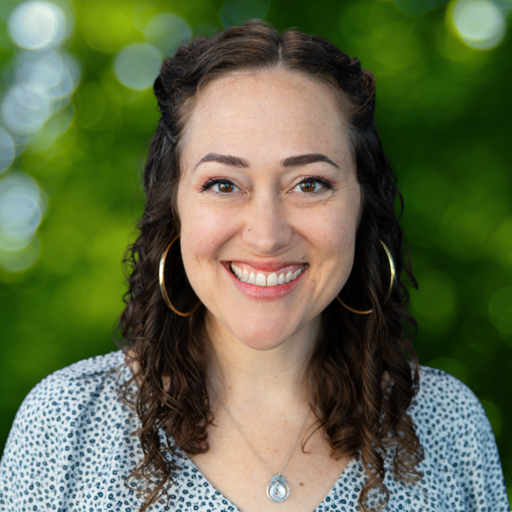 A woman with shoulder-length curly brown hair, smiling, wearing a patterned blouse and hoop earrings, standing in front of a background with dark green foliage.