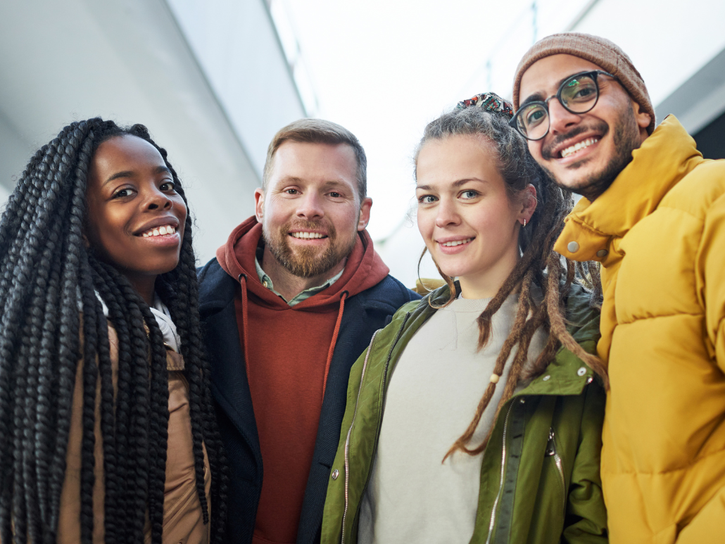 A group of four diverse young adults smiling together, wearing casual and winter clothing.