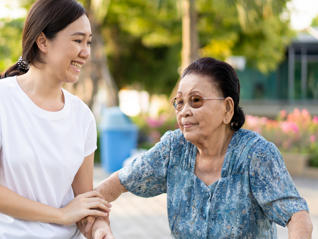 A young woman in a white shirt assisting an older woman with a walker in a park setting.