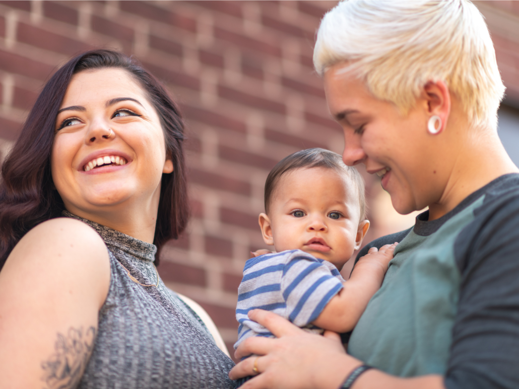 Smiling couple holding a baby outside in front of a brick wall.