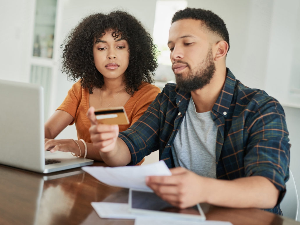 Two people sitting at a table with a laptop, one holding a credit card and the other reviewing documents.