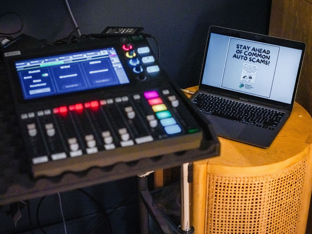 Audio mixer and laptop displaying "Stay Ahead of Common Auto Scams!" at the auto comic launch party.