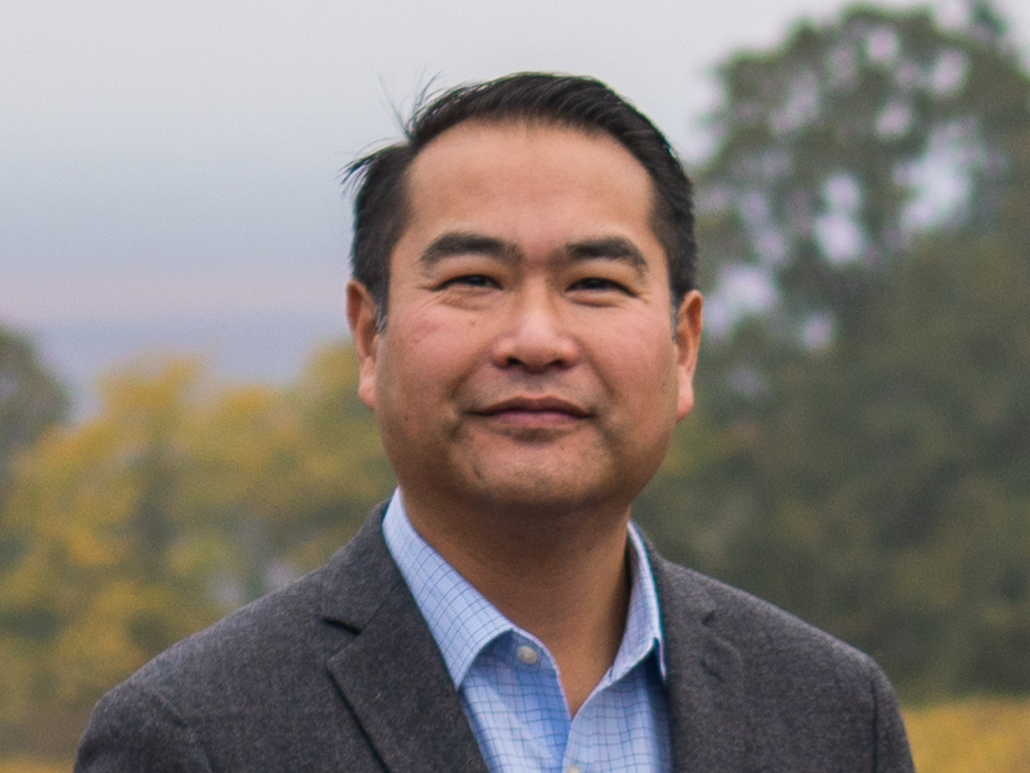 Portrait of Justin Baxter wearing a suit jacket and blue shirt, standing outdoors with trees in the background.