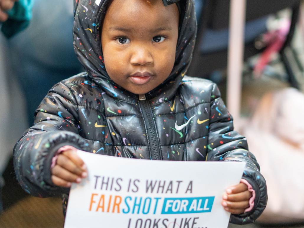 Young child wearing a patterned jacket holding a sign that reads 'This is what a fair shot for all looks like.