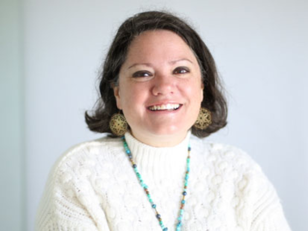 A woman with short dark hair, smiling, wearing a white sweater and turquoise necklace, standing in front of a white background.