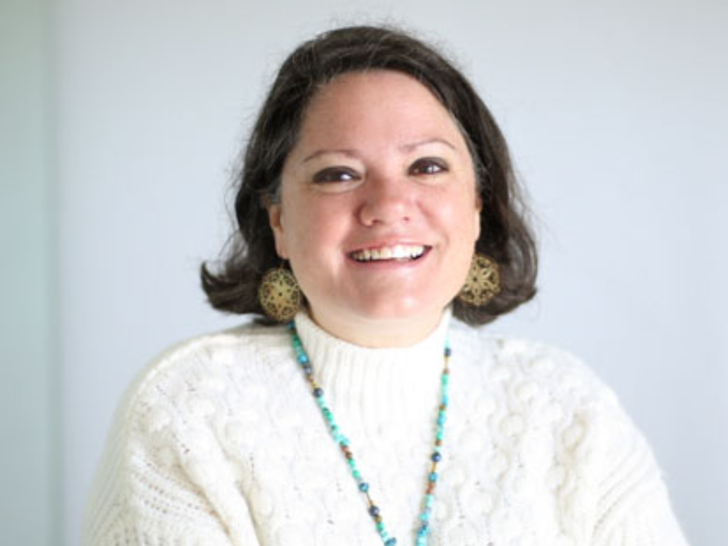   A woman with short dark hair, smiling, wearing a white sweater and turquoise necklace, standing in front of a white background.