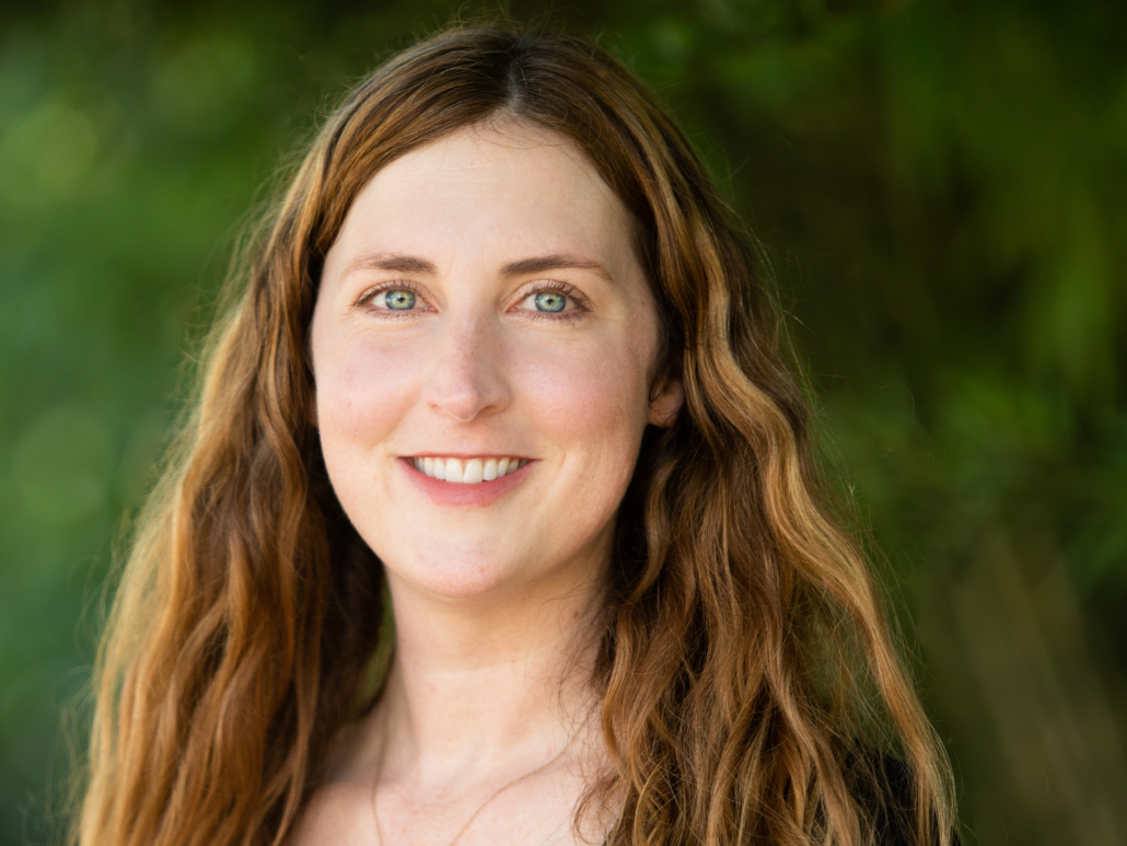 A woman with long reddish hair, smiling, wearing a black top, and standing in front of a green natural background.