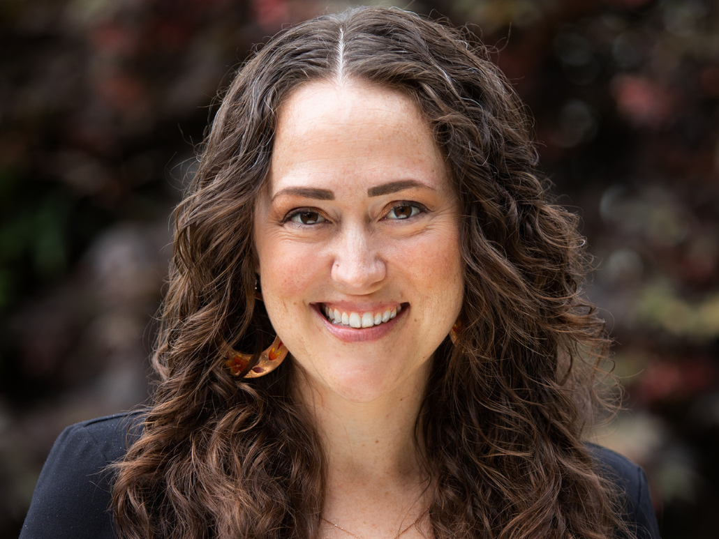 A woman with long curly brown hair, smiling, wearing a black top, standing in front of a background with colorful foliage.