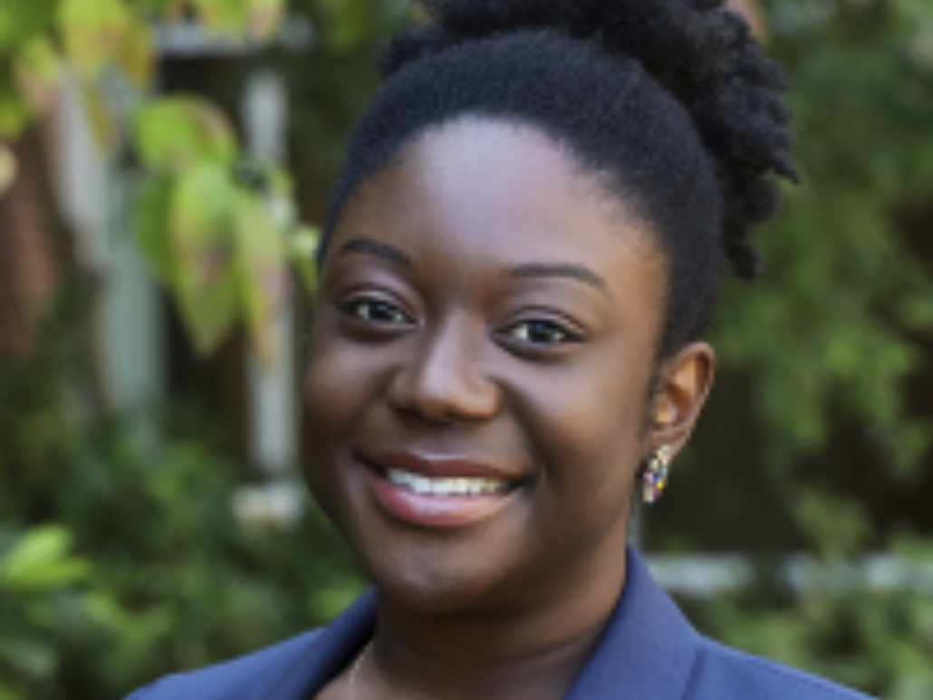 A close-up portrait of Angela, smiling with a blurred green foliage background.