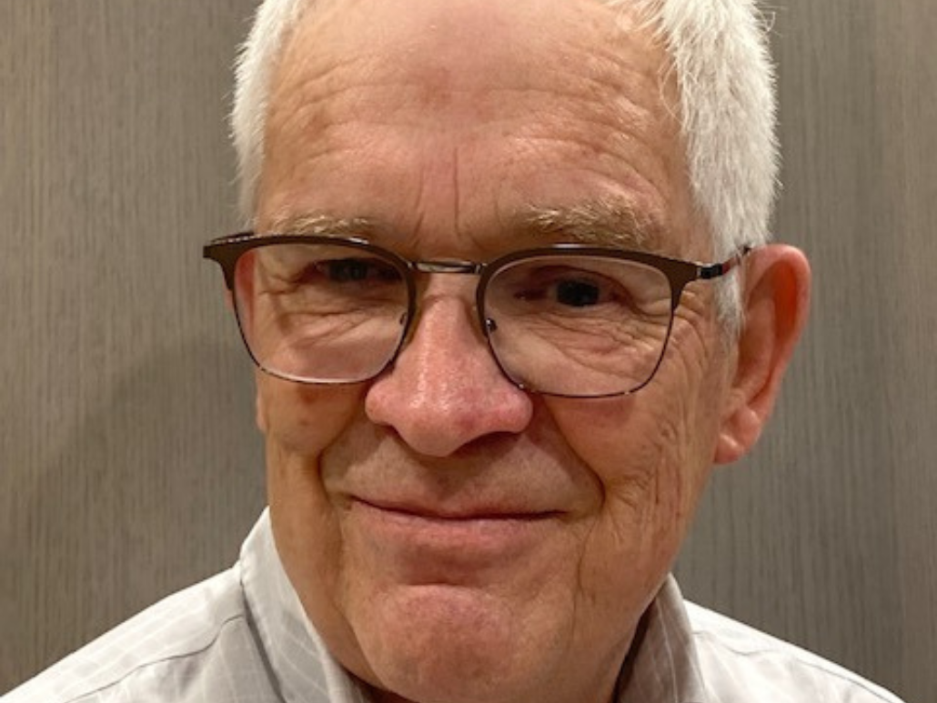 A close-up portrait of Dick, an older man with glasses, smiling with a neutral background.