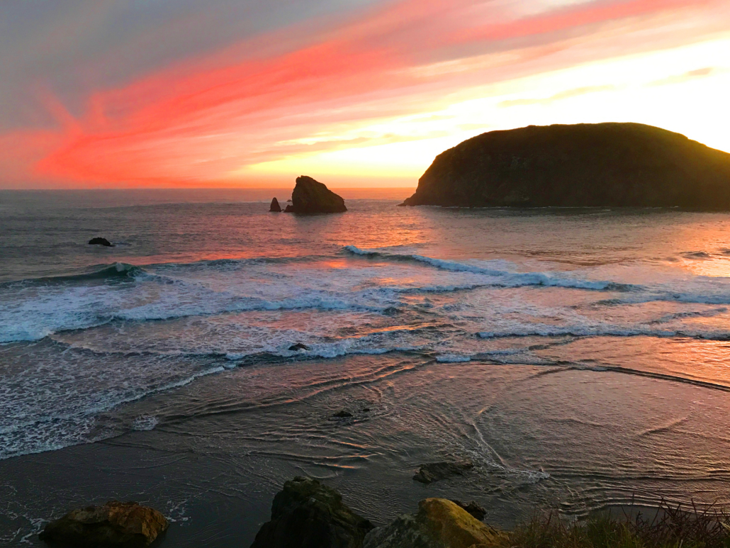 Scenic sunset at the beach with waves and a rock formation.