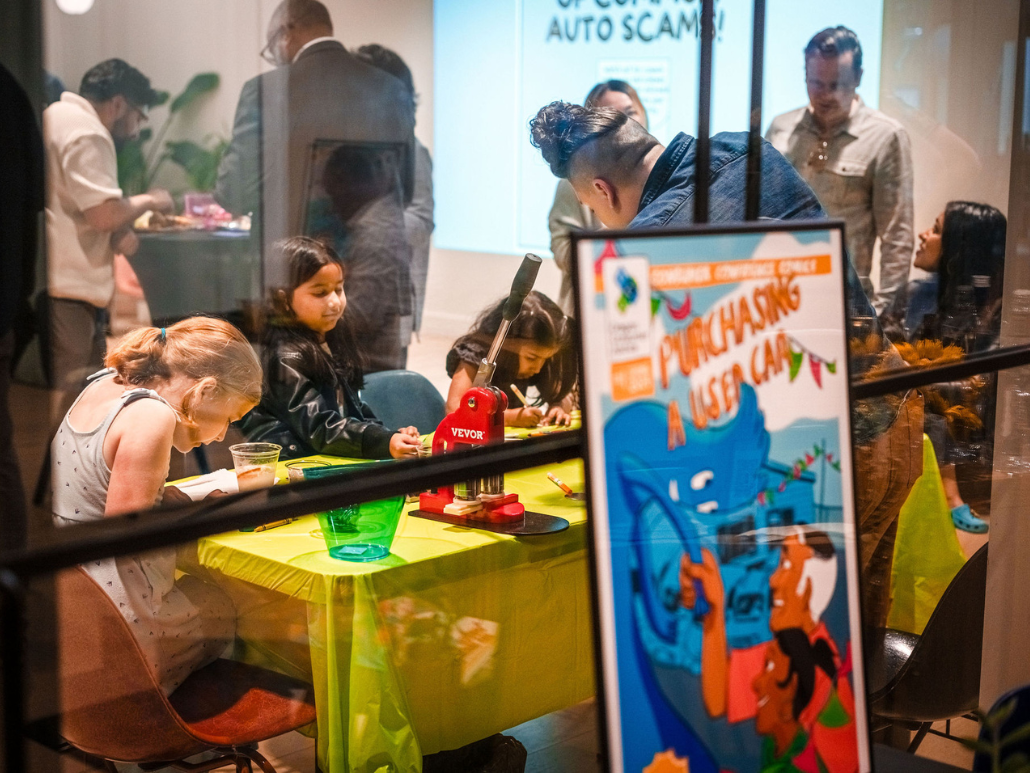 A group of children and adults are engaged in an activity at a community event, with a colorful poster titled "Purchasing a Used Car" visible in the foreground.