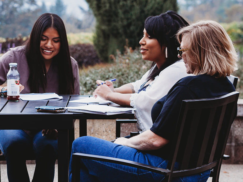 Three women sitting at a table outdoors, smiling and working on documents together.