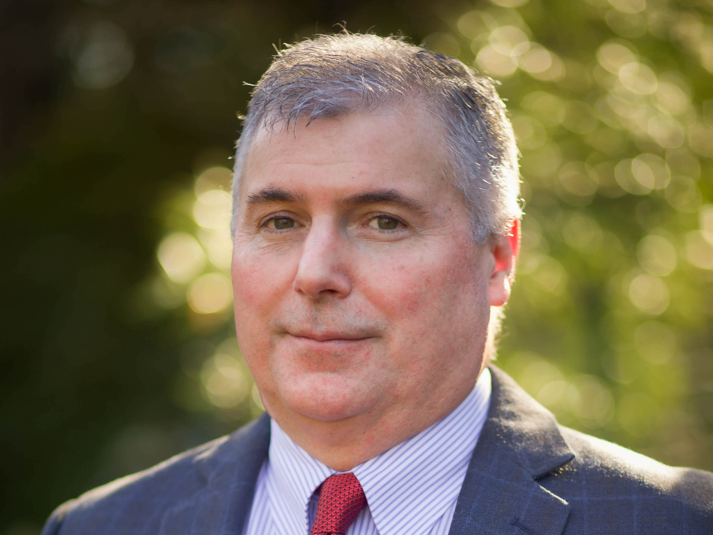 A man with short gray hair, wearing a suit, white shirt, and red tie, standing in front of a blurred natural background with bokeh effect.