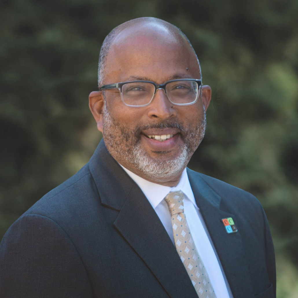 A man with a salt and pepper beard, and glasses, smiling, wearing a dark suit and light-colored tie, standing outdoors with greenery in the background.