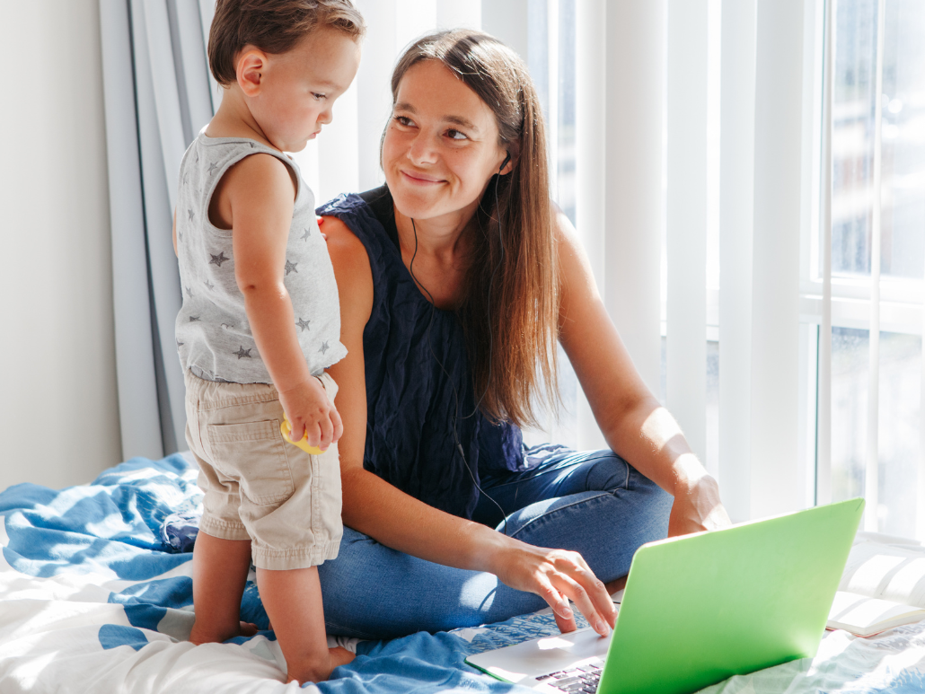 Woman sitting on a bed with a toddler, working on a laptop.