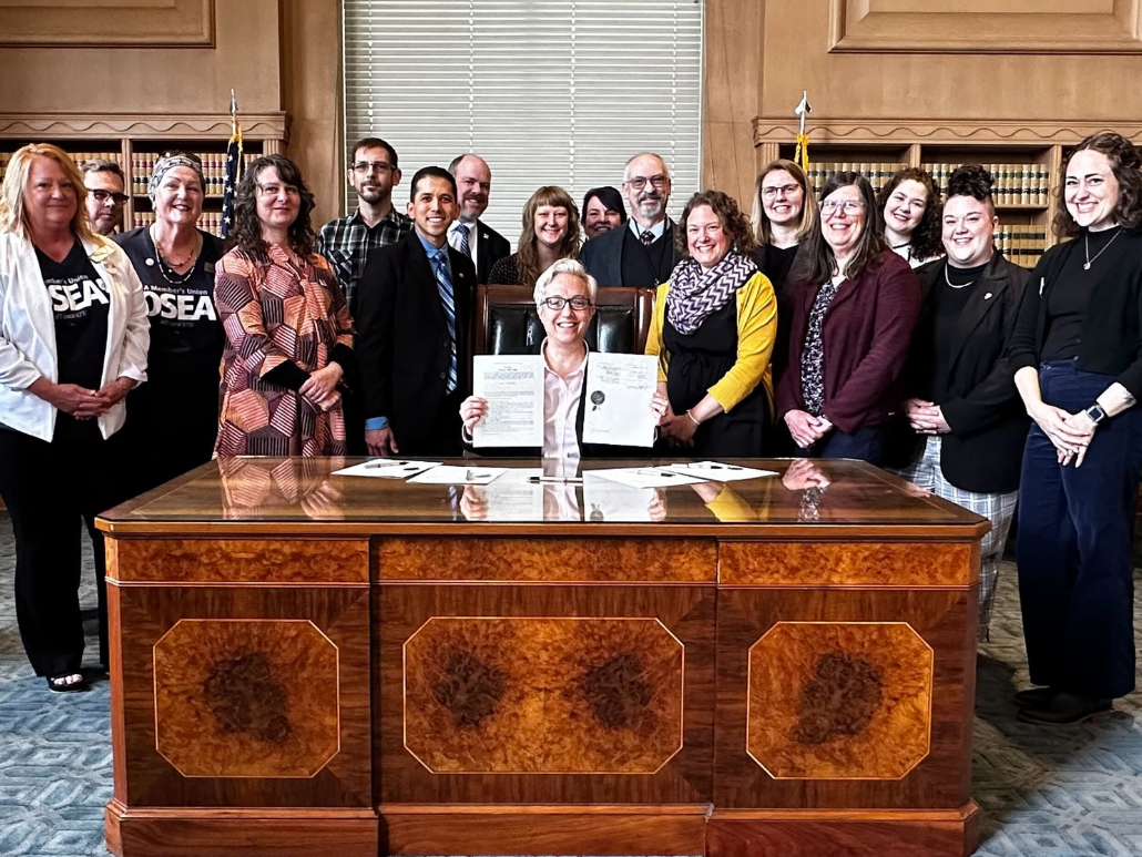 A group of advocates standing behind a large wooden desk, with Governor Tina Kotek, seated in the center holding up the ceremonially signed documents for SB 1595. They are in a formal room with flags and bookshelves in the background.