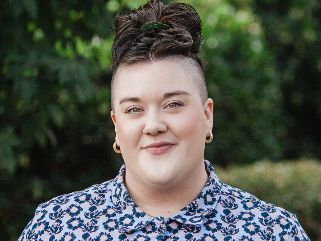 A person with short dark hair styled in a top knot, wearing gold hoop earrings and a floral patterned shirt, smiling in front of a green leafy background.