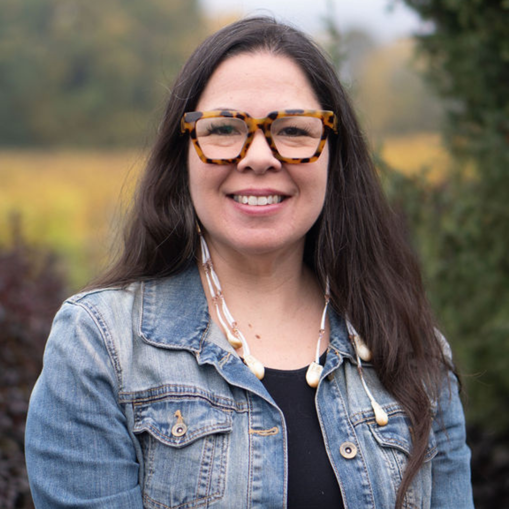 A woman with long dark hair, wearing tortoiseshell glasses, a denim jacket, and a necklace, smiling in an outdoor setting with a blurred background of trees and bushes.