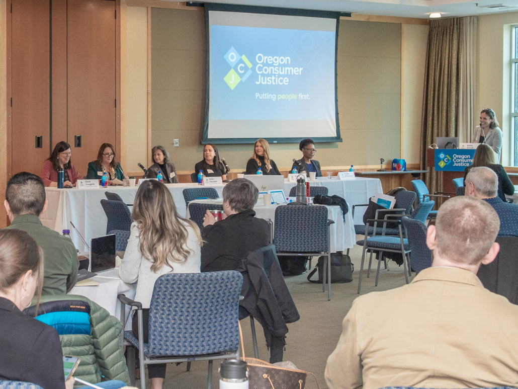 March 2023 CLE event hosted by OCJ featuring a panel of speakers at a long table in front of an audience, with a projector screen displaying "Oregon Consumer Justice" and the tagline "Putting people first."