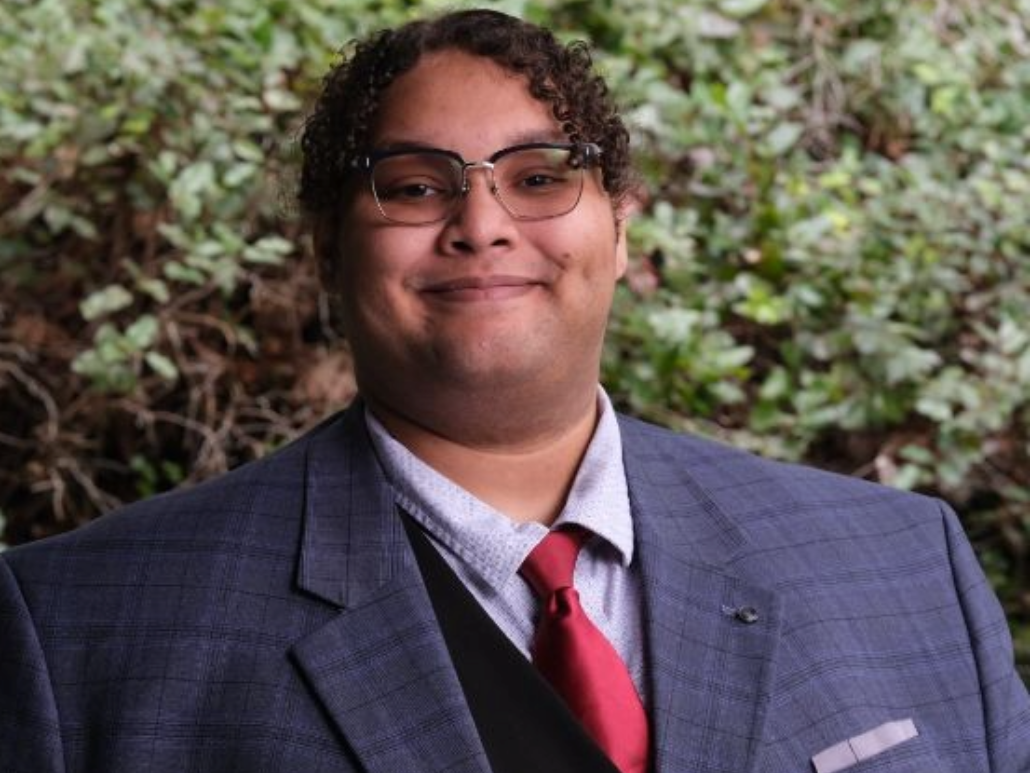 Portrait of Nik wearing a suit and tie, smiling outdoors with greenery in the background.