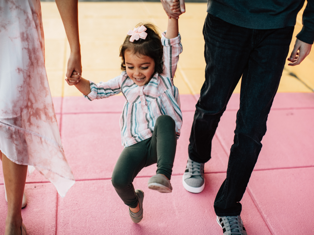 Niño pequeño con coletas tomados de la mano y balanceándose entre dos cuidadores.